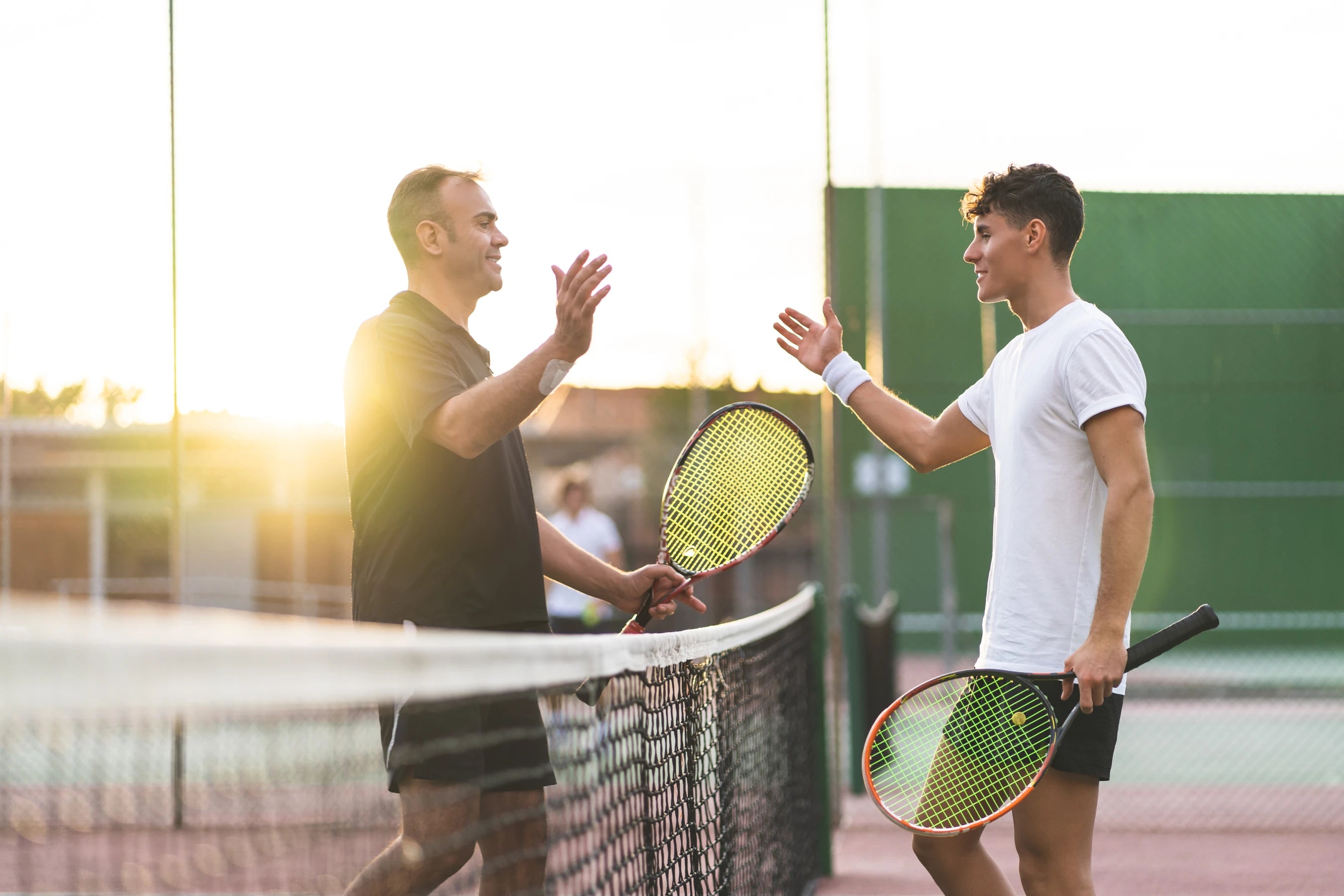 Isä ja aikuinen poika pelaamassa tennistä auringonlaskussa – elämä, johon energia riittää